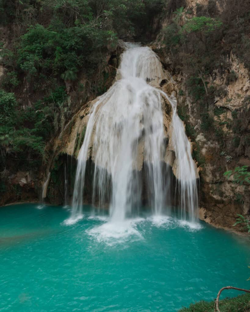 Cascadas El Chiflon: Without a Tour + Overnight Stay 25 Cascada Quinceanera is shown in full, with water pouring down from a high cliff into a bright turquoise pool. The surrounding cliffs and greenery frame the waterfall, creating a stunning natural vista.