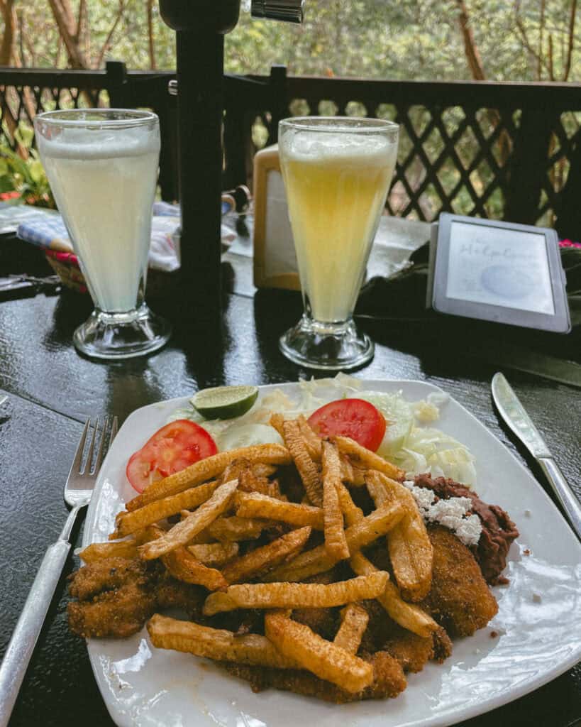 Cascadas El Chiflon: Without a Tour + Overnight Stay 28 A lunch meal of breaded fish fillets topped with French fries, accompanied by beans, lettuce, tomatoes, and lime slices. Two frothy drinks sit in tall glasses in the background at an outdoor restaurant.