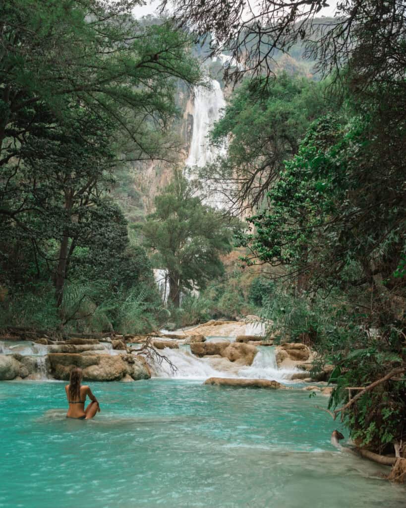 Cascadas El Chiflon: Without a Tour + Overnight Stay 8 Sally sits in the clear turquoise water of the river, surrounded by trees and with a distant view of a waterfall in the background, enjoying the peaceful scenery at Cascadas El Chiflón.
