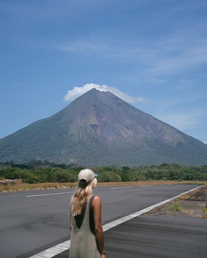 Sally Sees standing on a tarmac in Isla Ometepe, Nicaragua with a looming volcano behind her against a blue sky. 