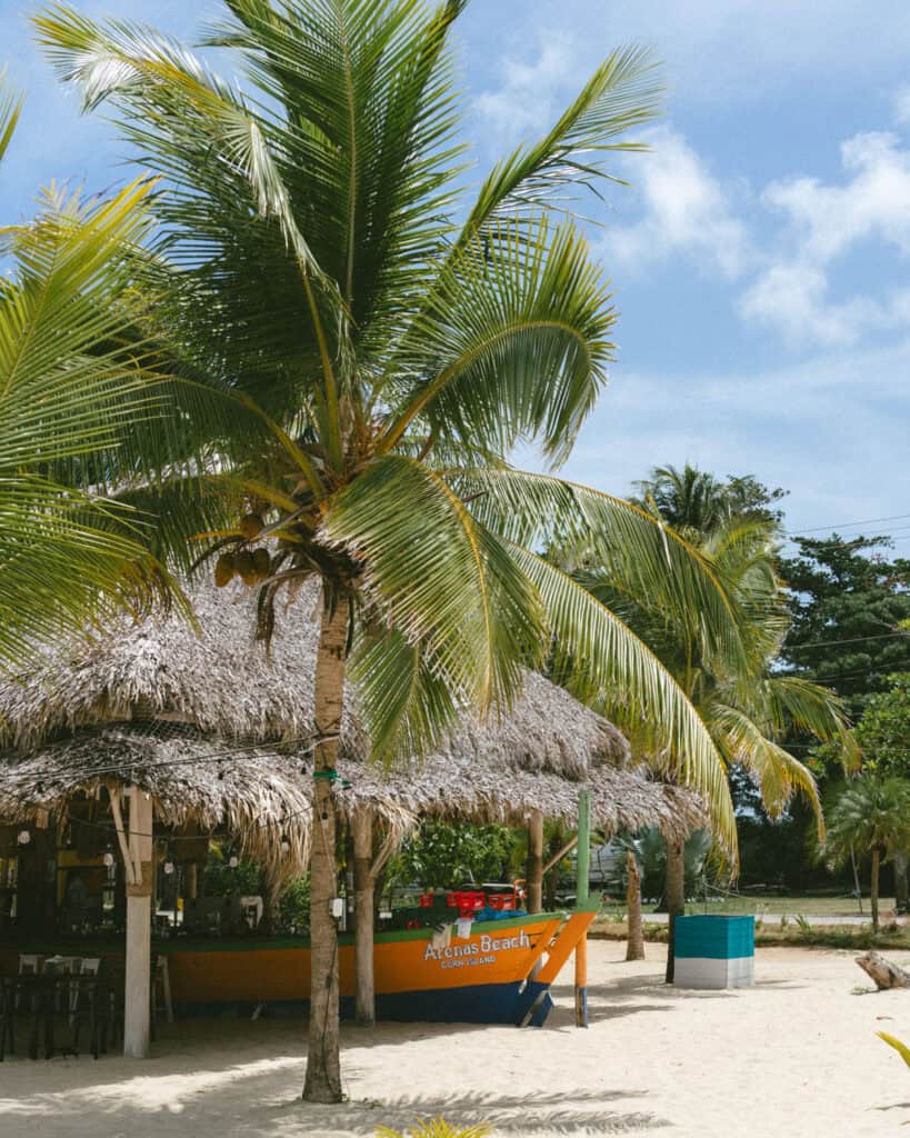 Big Corn Island, Nicaragua: The Underrated Corn Island 27 A bright yellow and blue boat labeled “Arenas Beach, Corn Island” resting beside a bar under a thatched roof, with palm trees providing shade.