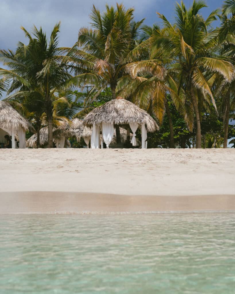 Big Corn Island, Nicaragua: The Underrated Corn Island 19 A scenic view from the ocean looking at Arenas Beach Club’s cabanas and palm trees, set against a backdrop of bright blue sky and sand.