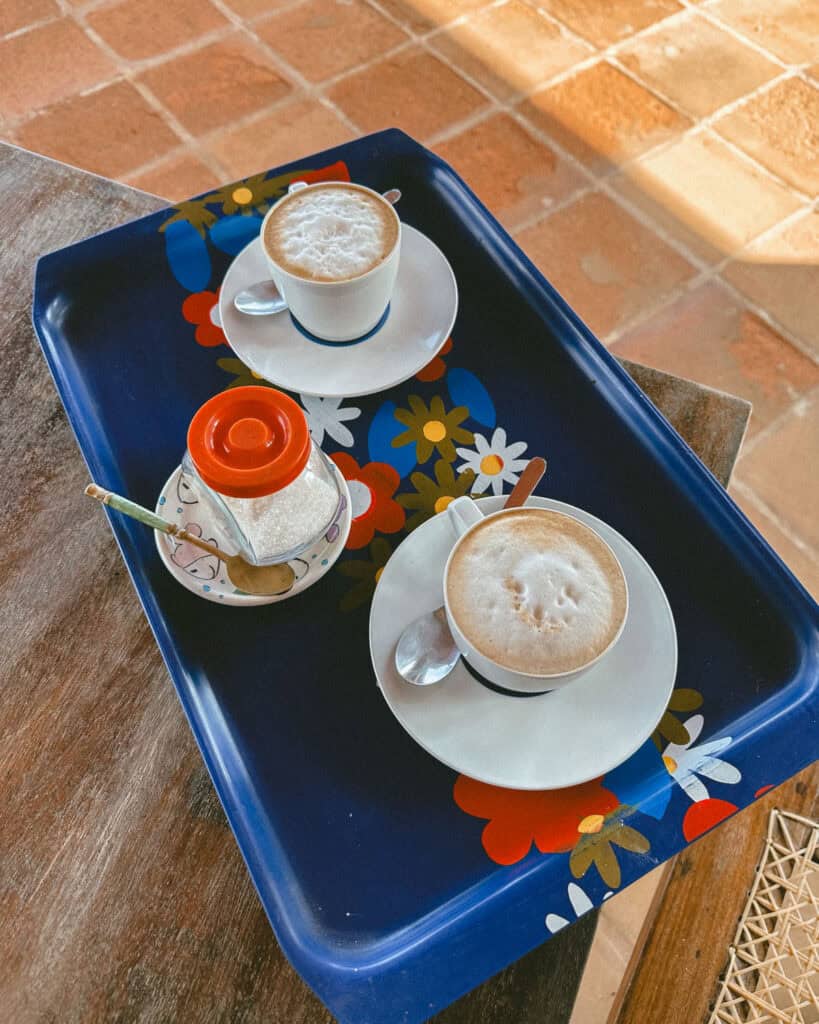 Big Corn Island, Nicaragua: The Underrated Corn Island 42 A tray with two frothy cups of coffee and sugar on a floral-patterned tray, set on a wooden table with terracotta tiles in the background.