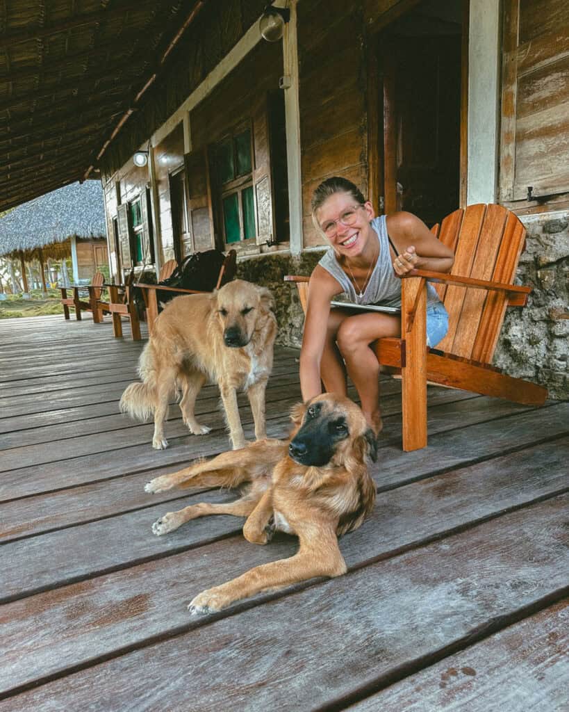 Big Corn Island, Nicaragua: The Underrated Corn Island 14 Sally sitting on the porch at La Princesa de la Isla, smiling while petting two dogs, with rustic wooden chairs and tropical scenery in the background.