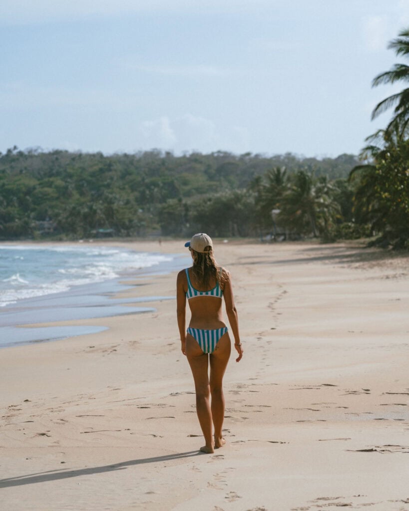 Big Corn Island, Nicaragua: The Underrated Corn Island 37 Sally walking along the edge of Long Bay Beach, wearing a striped bikini, with lush green hills in the distance.