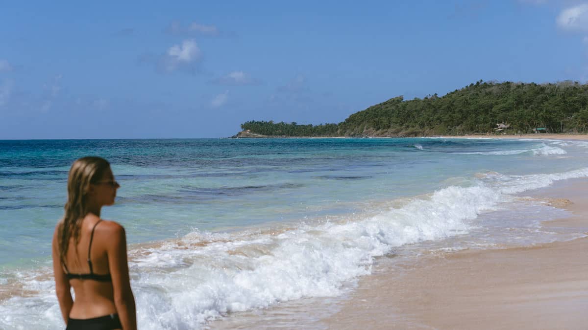 Big Corn Island, Nicaragua: The Underrated Corn Island 4 Sally standing at the waters edge on Long Bay Beach, Big Corn Island. She is out of focus in the foreground with the turquoise waters and green forested hills clear in the background.