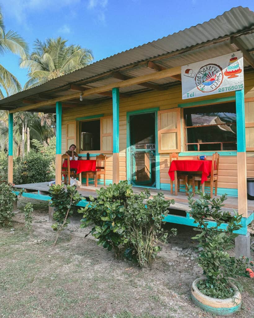 Big Corn Island, Nicaragua: The Underrated Corn Island 43 The exterior of Island Bakery and Sweets on Big Corn Island, featuring a wooden building with turquoise trim and red tablecloths on the porch. Sally is sitting and smiling at one of the tables.