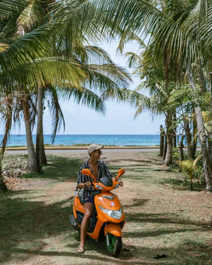 Big Corn Island, Nicaragua: The Underrated Corn Island 39 Brayden seated on his orange scooter beneath towering palm trees, with the ocean visible through the trees on Big Corn Island.