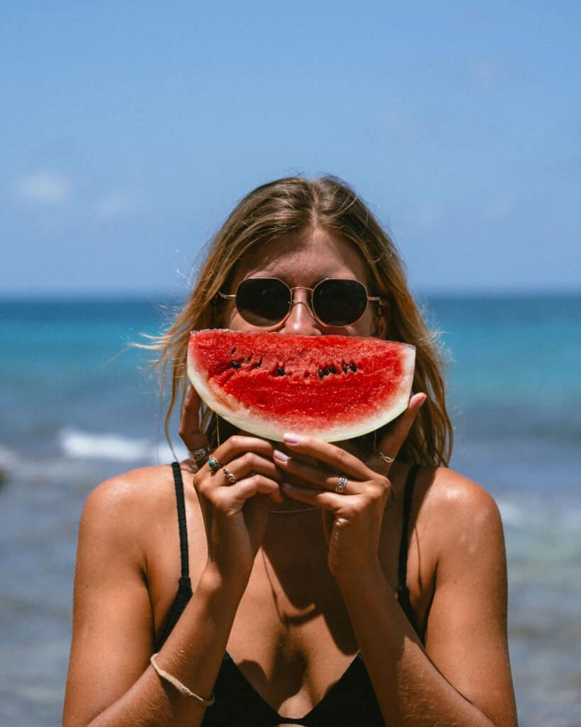 Big Corn Island, Nicaragua: The Underrated Corn Island 48 Sally holding a bright slice of watermelon up to her face, wearing sunglasses, with a vibrant blue ocean horizon behind her on Big Corn Island.