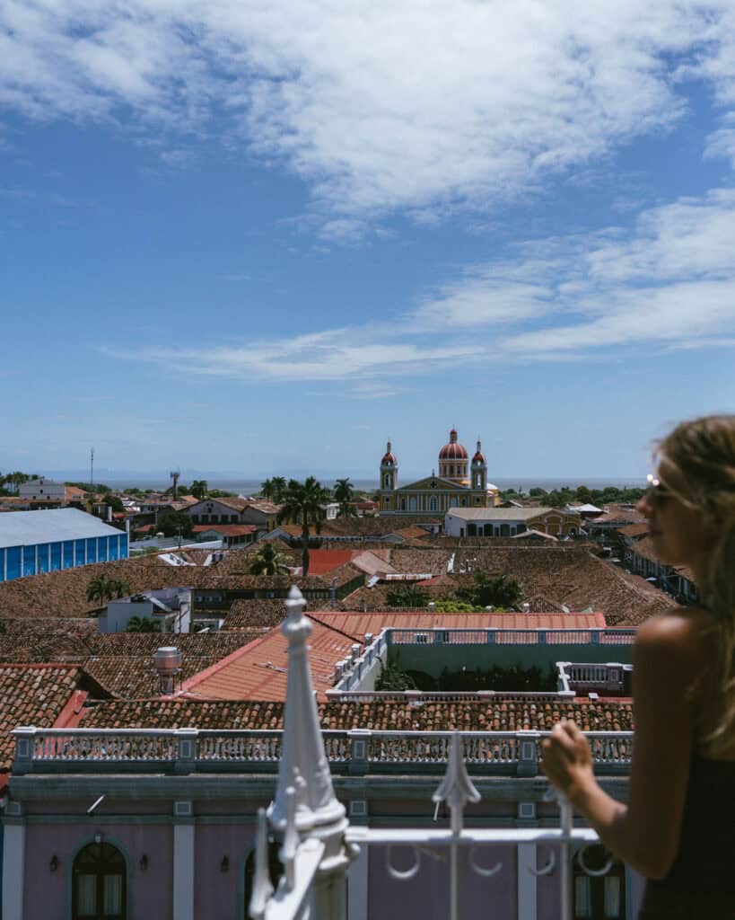 Granada, Nicaragua Travel Guide: Our Honest Thoughts 16 Sally stands on the balcony of the Iglesia de la Merced bell tower, overlooking Granada, Nicaragua, with the iconic red-domed Cathedral of Granada visible in the distance under a clear blue sky.