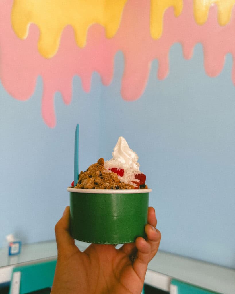 Granada, Nicaragua Travel Guide: Our Honest Thoughts 26 A hand holding a green cup filled with creamy frozen yogurt, topped with cookie crumbles, cherries, and coconut, against a pastel wall with colorful drips.