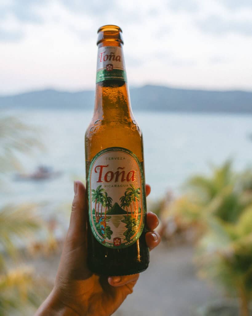 Nicaragua Travel Guide: Land of Lakes and Volcanoes 49 A hand holding a chilled bottle of Toña beer with the serene waters of Laguna de Apoyo in the background, framed by lush greenery and distant mountains.