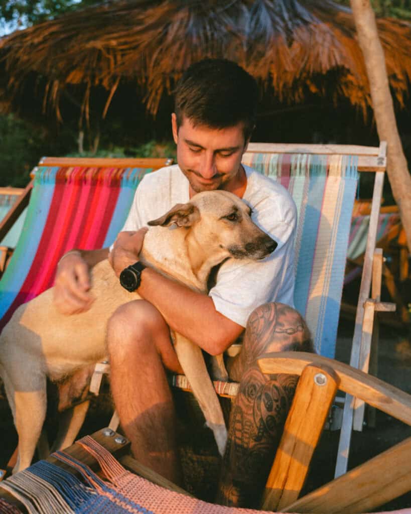 Laguna de Apoyo, Nicaragua Travel Guide (Stay > Day) 15 Brayden sitting on a colorful hammock chair under a thatched roof, affectionately holding a dog, with the warm morning light at Laguna de Apoyo.