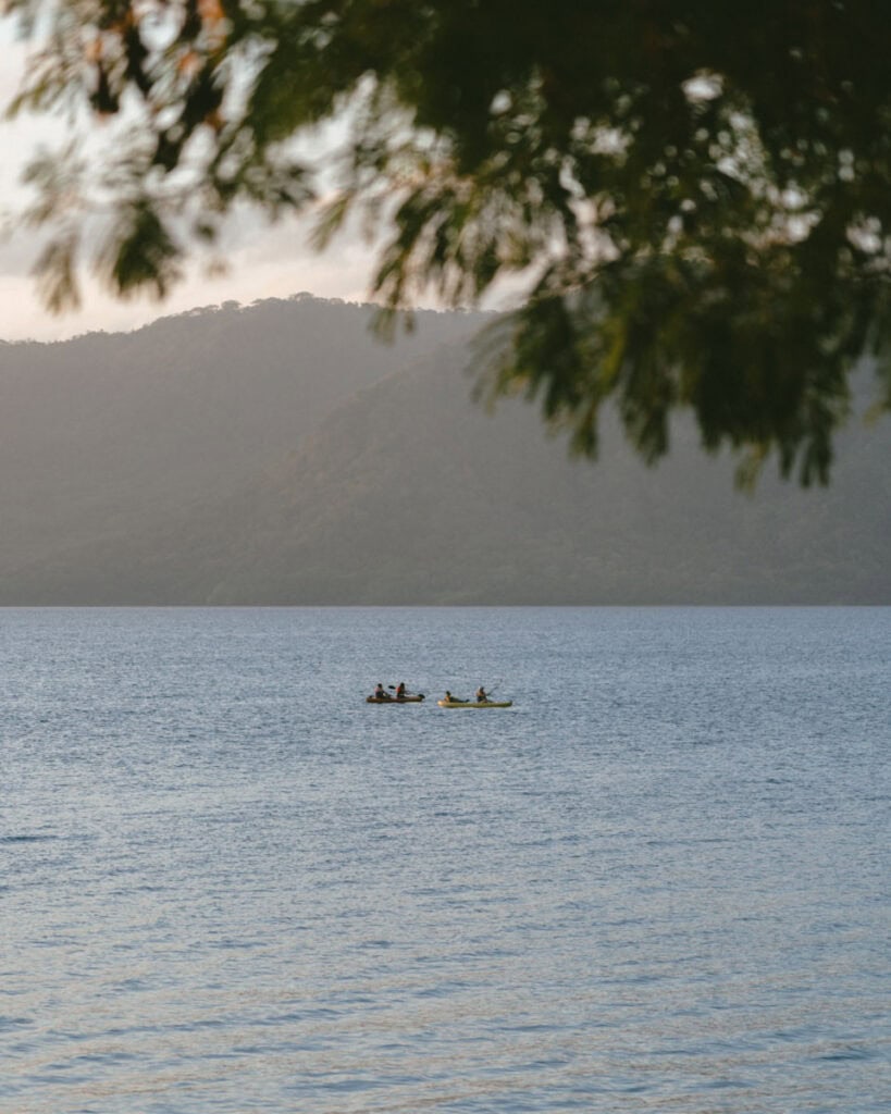 Laguna de Apoyo, Nicaragua Travel Guide (Stay > Day) 13 A group of people kayaking on the calm waters of Laguna de Apoyo, with a backdrop of lush green hills and soft morning light.