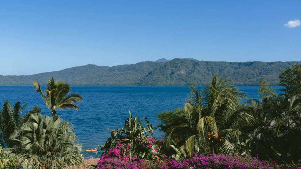 Laguna de Apoyo, Nicaragua Travel Guide (Stay > Day) 7 A scenic shot of the lake at Laguna de Apoyo, framed by tropical foliage and purple flowers in the foreground, with rolling hills in the distance.