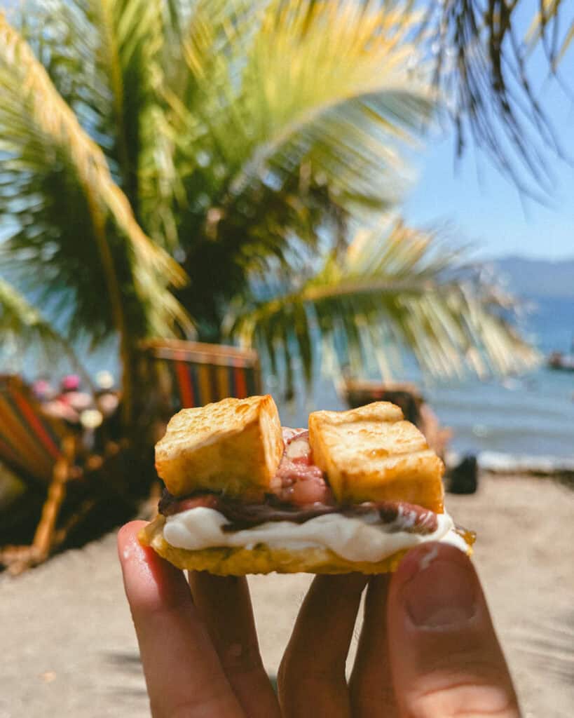 Nicaragua Travel Guide: Land of Lakes and Volcanoes 50 A hand holding a small snack of crispy fried plantain topped with cream and beans, with a view of Laguna de Apoyo and lounge chairs beneath palm trees.