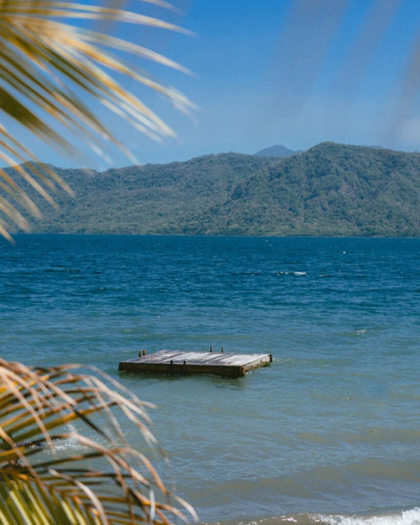 Laguna de Apoyo, Nicaragua Travel Guide (Stay > Day) 8 A floating wooden pontoon seen through swaying palm leaves, with the tranquil Laguna de Apoyo and distant hills under a clear sky.