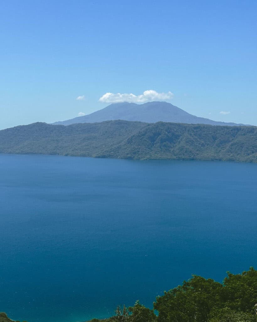 Laguna de Apoyo, Nicaragua Travel Guide (Stay > Day) 17 Laguna de Apoyo from an elevated vantage point, showcasing the expansive blue crater lake surrounded by verdant forest, with Mombacho Volcano in the background.