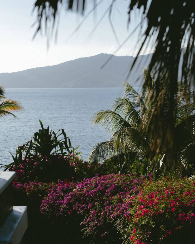 Laguna de Apoyo, Nicaragua Travel Guide (Stay > Day) 11 A view towards Laguna de Apoyo in soft morning light with flowers and palm trees in the foreground.