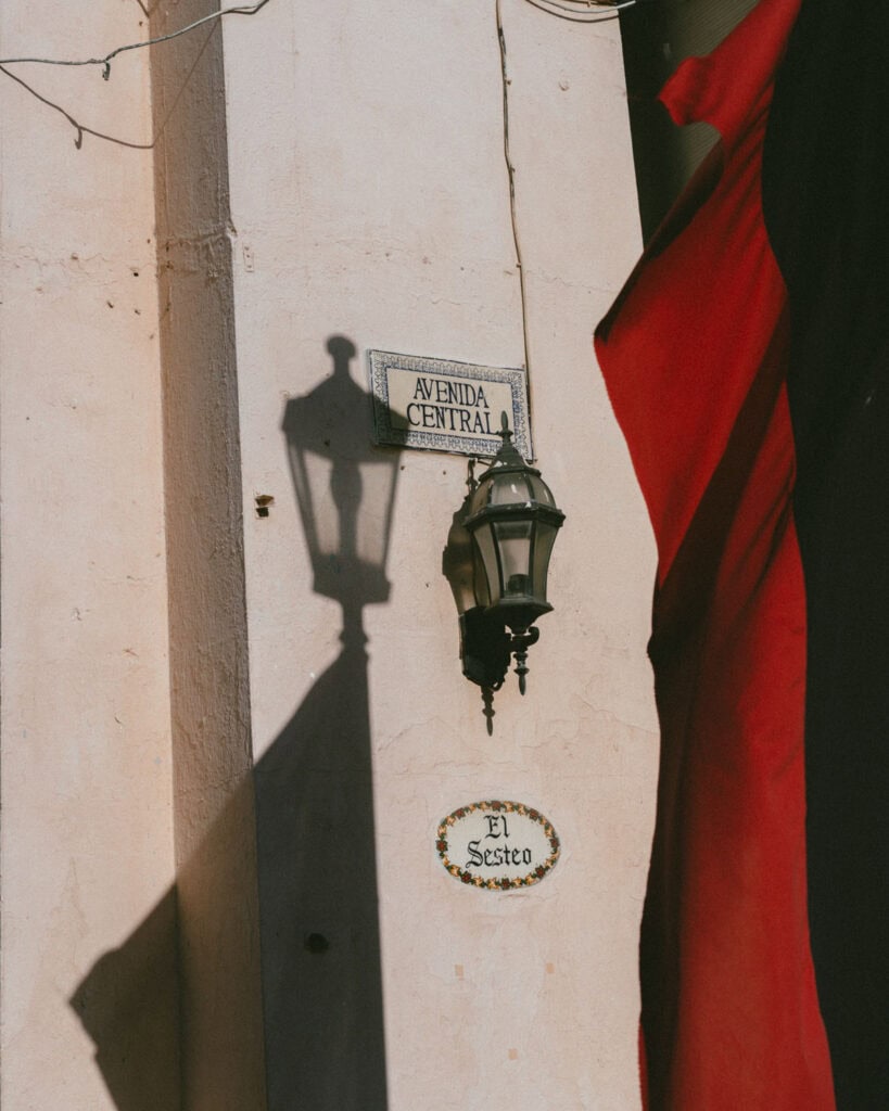 18 Best Things to Do in Leon, Nicaragua 39 A close-up of a vintage lamp casting a shadow on a stucco wall in León, Nicaragua, with a tiled sign reading "Avenida Central" and a plaque below it labeled "El Sesteo."
