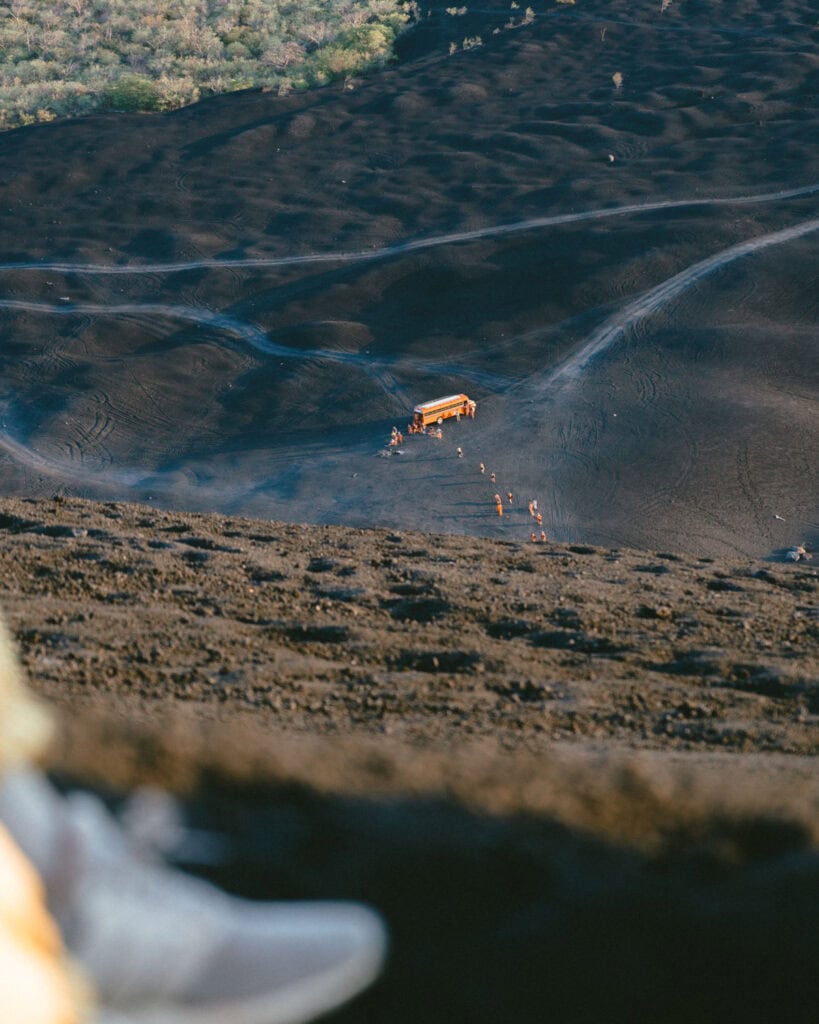 Epic Volcano Boarding in Nicaragua at Cerro Negro Volcano 8 An orange "Bigfoot" bus parked at the base of Cerro Negro volcano on the black volcanic rock, with tourists preparing for their volcano boarding adventure.