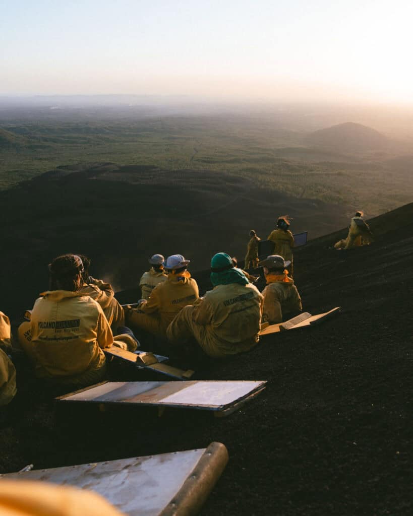 Epic Volcano Boarding in Nicaragua at Cerro Negro Volcano 20 A group of adventurers dressed in yellow volcano boarding suits, seated on the volcanic slope of Cerro Negro, enjoying the expansive view at sunset.