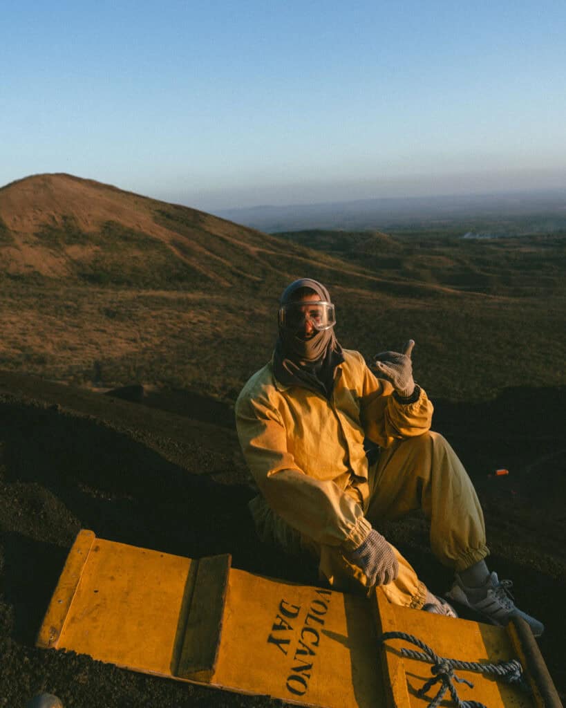 Epic Volcano Boarding in Nicaragua at Cerro Negro Volcano 5 Brayden in full volcano boarding gear sitting on the black volcanic slope, holding his board and giving a shaka.