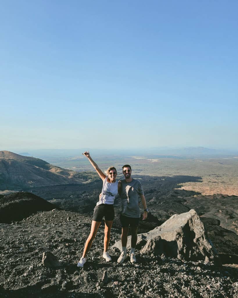 Epic Volcano Boarding in Nicaragua at Cerro Negro Volcano 12 Sally and Brayden standing on a volcanic slope in Cerro Negro, Leon, Nicaragua. They are dressed in casual hiking attire, enjoying the expansive view under a bright blue sky.