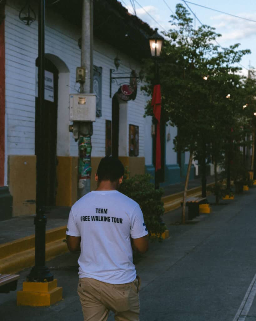 18 Best Things to Do in Leon, Nicaragua 8 A man wearing a "Team Free Walking Tour" t-shirt strolls down a picturesque street in León, Nicaragua, lined with colorful colonial buildings and lamp posts.