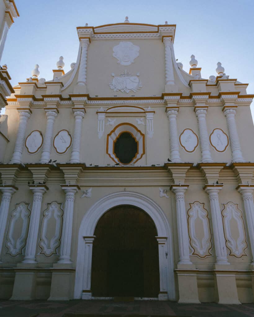18 Best Things to Do in Leon, Nicaragua 36 The front of Iglesia de La Merced in León, Nicaragua, with its soft pastel yellow tones and maroon and white skirts, highlighting its ornate architecture against a clear blue sky.