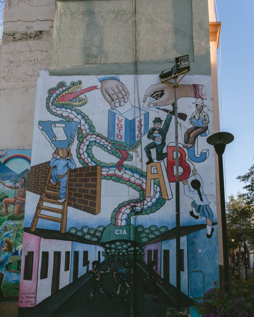 18 Best Things to Do in Leon, Nicaragua 21 A vivid mural in León, Nicaragua, depicting historical and cultural symbols, including a CIA sign, a ballot box labeled "Voto" and a serpent intertwined with figures.