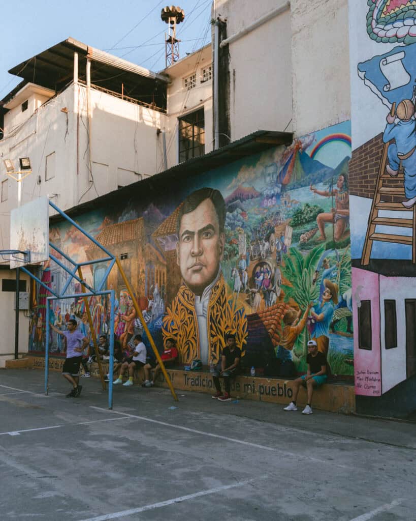 18 Best Things to Do in Leon, Nicaragua 23 A colorful mural showing a prominent historical figure surrounded by scenes of daily life and cultural heritage in León, Nicaragua. The mural is in the city's basketball court and local guys are sitting along a ledge in front of the wall.