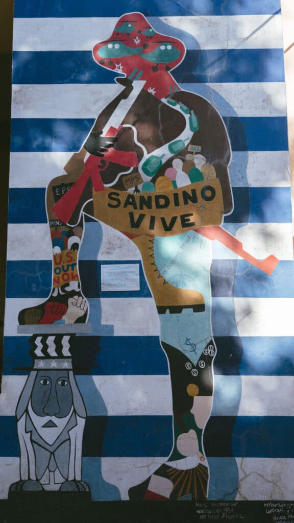18 Best Things to Do in Leon, Nicaragua 20 A mural on a blue and white striped wall, featuring a large figure with the text "Sandino Vive," accompanied by various symbolic elements, in León, Nicaragua.