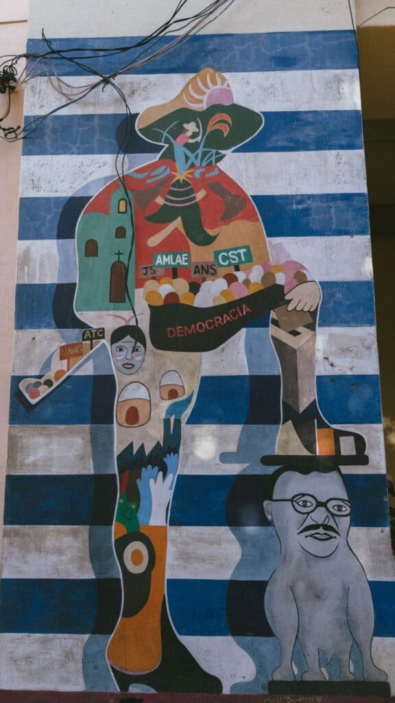 18 Best Things to Do in Leon, Nicaragua 19 A mural on a blue and white striped wall, featuring a large figure with its foot squashing a political opponent, in León, Nicaragua.