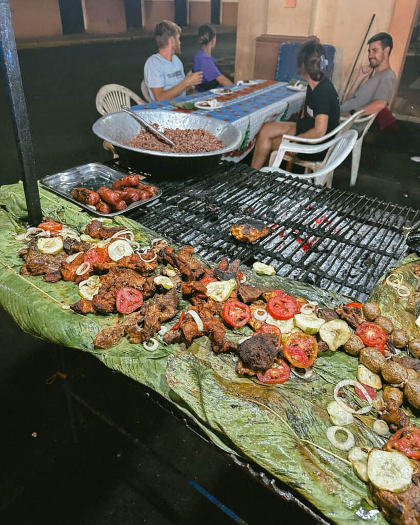18 Best Things to Do in Leon, Nicaragua 27 A lively evening at a local fritanga in León, Nicaragua, with sizzling grilled meats on a wood-fired BBQ and patrons seated at a street-side table.