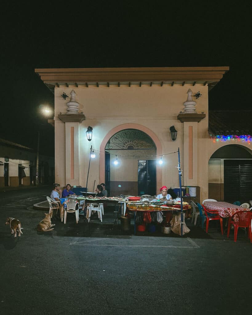 Epic Volcano Boarding in Nicaragua at Cerro Negro Volcano 26 A typical fritanga set up on a street in Leon at night, with a dimly lit BBQ and plastic chairs and tables with patrons eating, and a few dogs waiting for scraps.