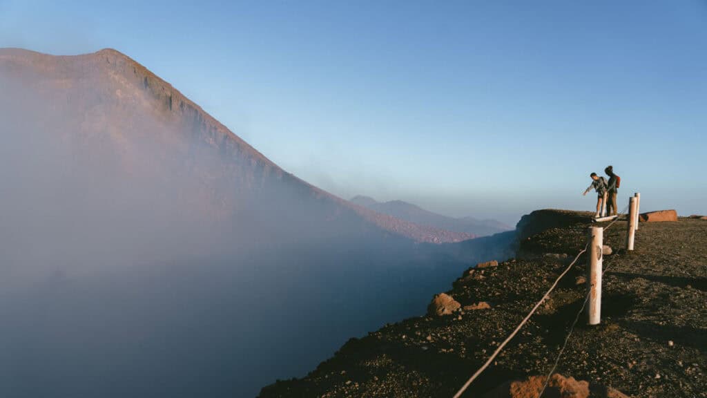 Hiking Telica Volcano in Nicaragua: A Magical Sunrise 13 Brayden and another figure standing near the crater edge, framed by a dramatic landscape of rising smoke and volcanic terrain. Brayden is pointing down into the crater.