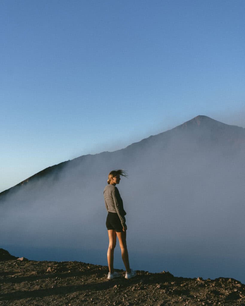 18 Best Things to Do in Leon, Nicaragua 45 Sally standing on the crater’s edge, her hair blowing in the wind, with smoke rising from Telica Volcano and a mountain backdrop.