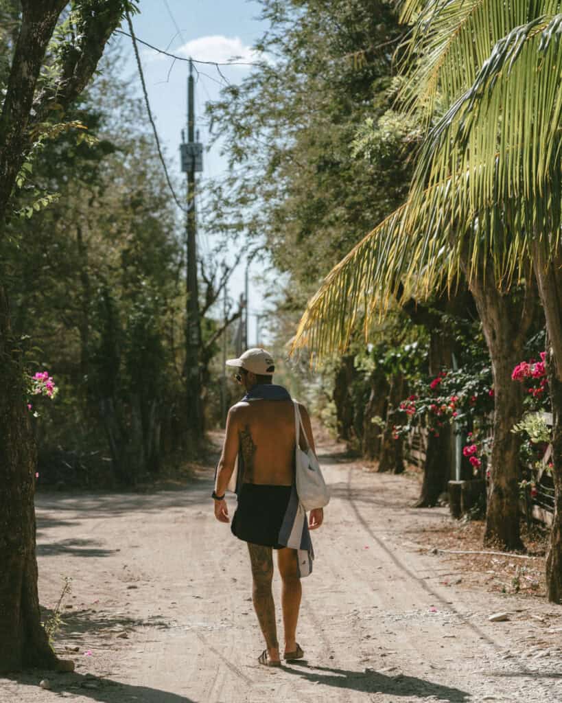 Popoyo, Nicaragua Travel Guide: Surf, Sand and Sunsets 6 Brayden walking shirtless on a dusty road in Popoyo, Nicaragua, surrounded by tropical foliage and vibrant flowers.