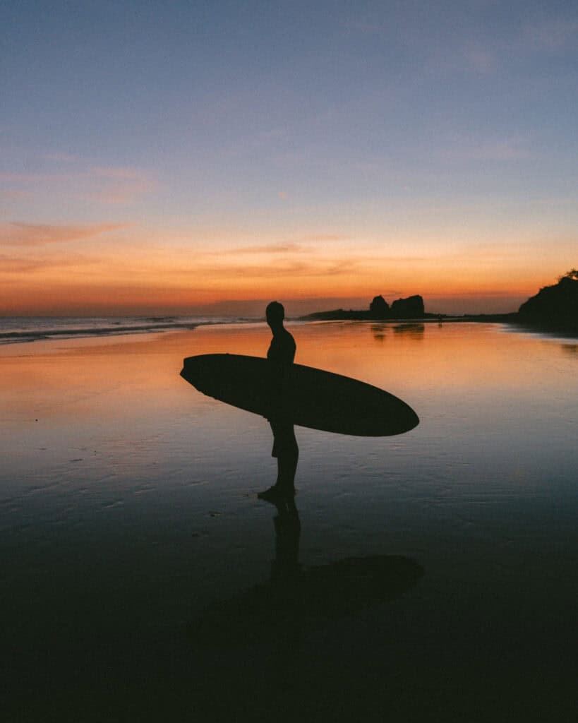 Popoyo, Nicaragua Travel Guide: Surf, Sand and Sunsets 3 Silhouette of Brayden holding a surfboard on Popoyo Beach at sunset, with vivid orange and pink hues reflecting on the wet sand, and the famous rock formations in the distance.