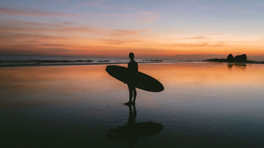 Popoyo, Nicaragua Travel Guide: Surf, Sand and Sunsets 21 Wide view of Brayden holding a surfboard at Popoyo Beach during sunset, with the horizon blending into warm tones.