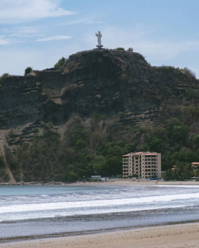 San Juan del Sur, Nicaragua Travel Guide: Worth the Hype? 4 A distant view of the Cristo de La Divina Misericordia statue atop a hill, with waves gently rolling onto the beach below in San Juan del Sur, Nicaragua.