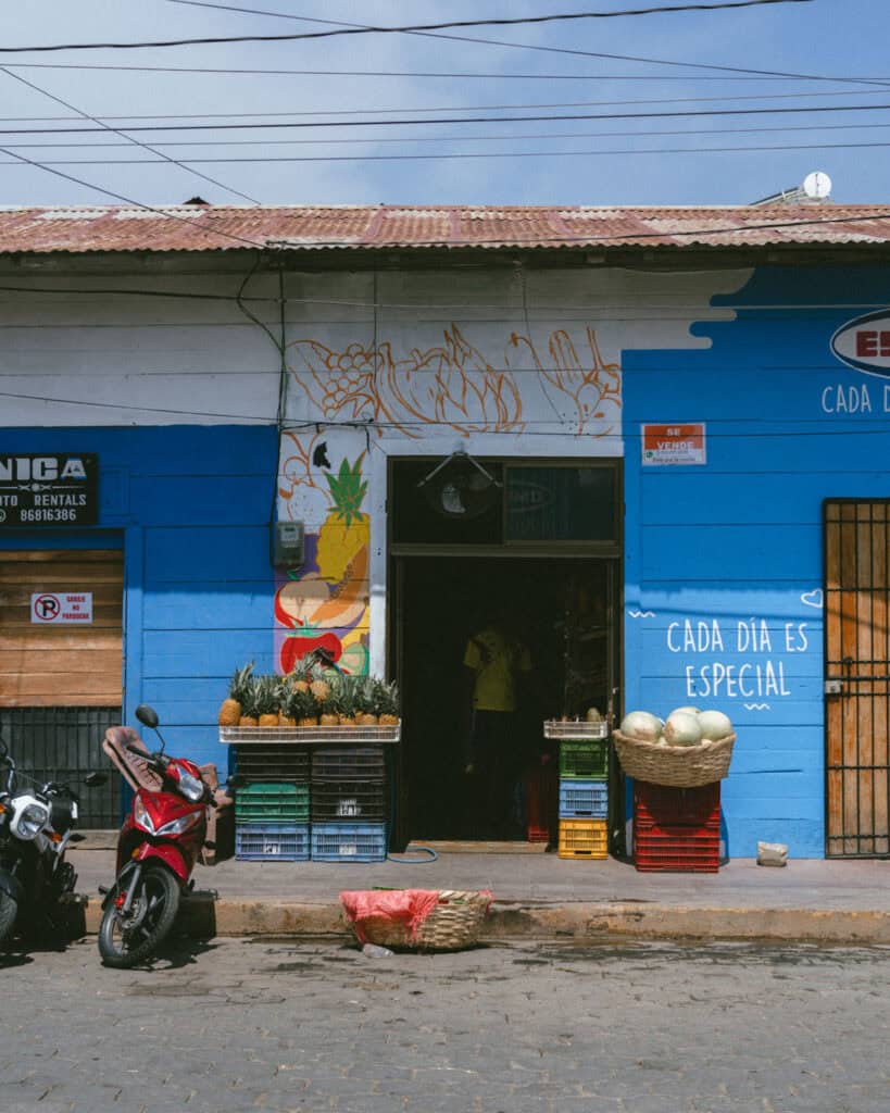 San Juan del Sur, Nicaragua Travel Guide: Worth the Hype? 5 A small fruit shop in San Juan del Sur, Nicaragua, with a vibrant blue facade, colorful fruit mural, and baskets of pineapples and melons outside.