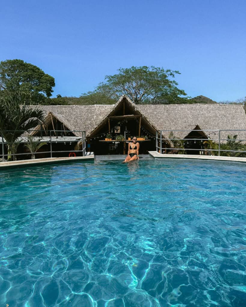 San Juan del Sur, Nicaragua Travel Guide: Worth the Hype? 26 Sally sitting on the edge of the TreeCasa pool, with the resort’s iconic thatched-roof structures and vibrant greenery in the background.