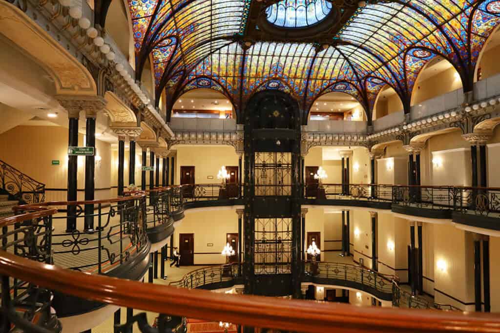 Ultimate Guide to Centro Historico, Mexico City 28 Ornate historic interior in Gran Hotel Ciudad de México Zócalo View Mexico City featuring a grand multi-level gallery with curved wrought-iron balconies, a central vintage elevator, and a stunning stained-glass dome ceiling in warm gold, blue, and red tones illuminating the elegant atrium.