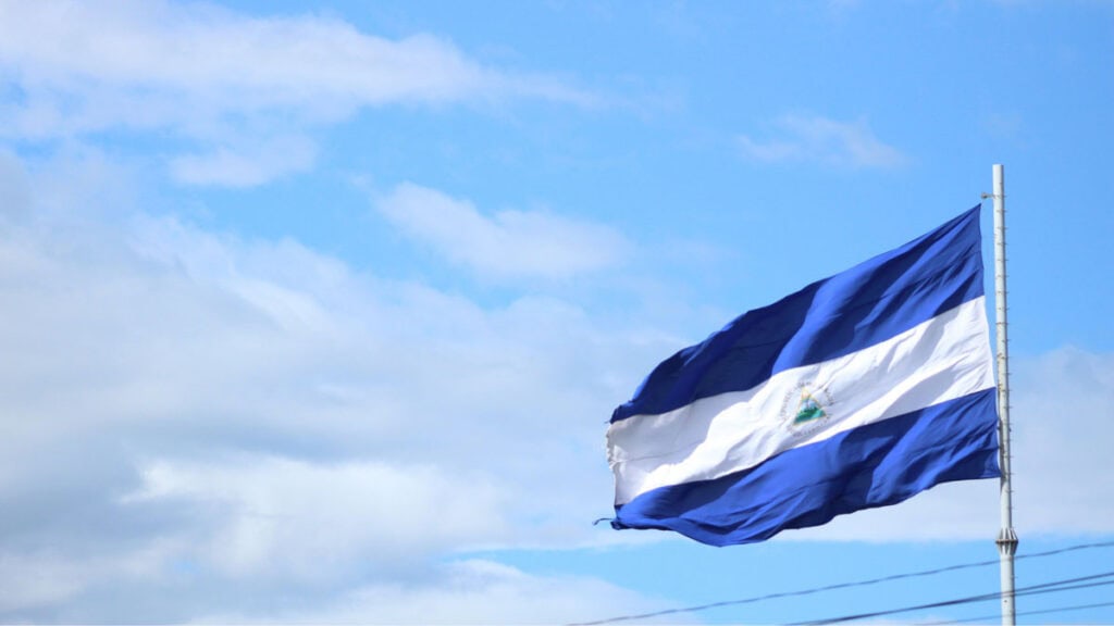 Nicaragua Travel Guide: Land of Lakes and Volcanoes 31 The Nicaraguan flag, with two outer blue horizontal stripes with a white strip in the middle and a triangle icon, on a flagpole flapping in the freeze against a blue, slightly cloudy sky.