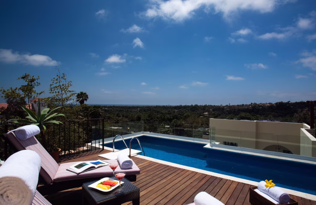 Where to Stay in Todos Santos, Mexico: Hotel & Area Guide 16 A luxurious rooftop pool in Guaycura Boutique Spa Todos Santos overlooking lush greenery and a distant coastline, with striped lounge chairs, rolled towels, fresh fruit, and cocktails set for a relaxing day under a bright blue sky.