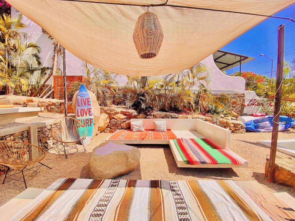 Where to Stay in Todos Santos, Mexico: Hotel & Area Guide 17 A vibrant and cozy surf hostel patio in Baja, decorated with colorful Mexican blankets, lounge seating, woven chairs, and a surfboard sign that reads “Live Love Surf.”