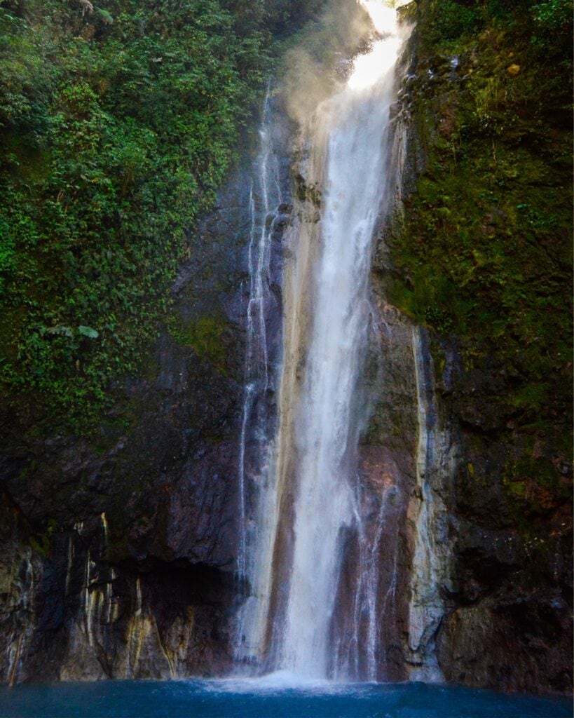 Bajos del Toro Travel Guide: Costa Rica's Waterfall Wonderland 14 A tall waterfall in Bajos del Toro, Costa Rica, tumbling down a cliff face surrounded by greenery into a small blue pool below. The waterfall is narrow and tall, not curtain-like.