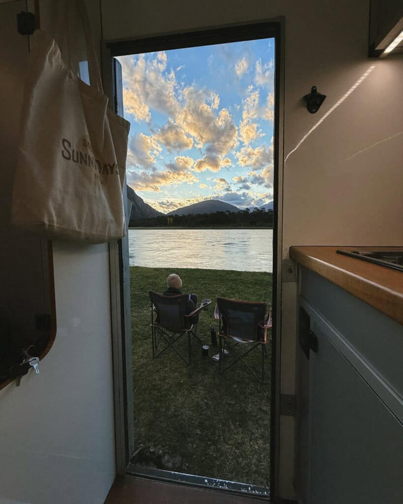 View from inside the camper van showing Brayden sitting by Río Baker in Caleta Tortel, Chile, facing a glowing sky filled with golden clouds at sunset
